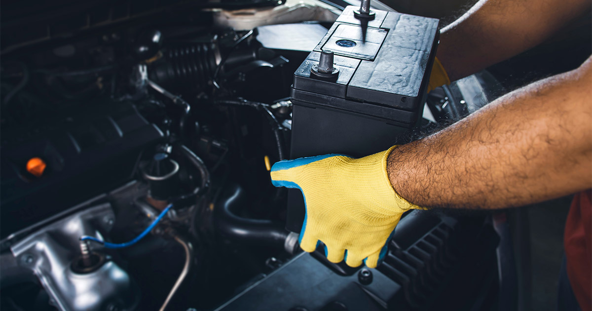 Person wearing yellow gloves removes a car battery from an engine bay. The image conveys maintenance and care in an automotive context.
