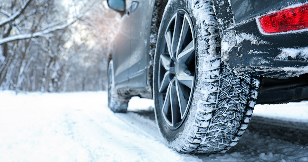 Close-up of a car tire with snow chains, driving on a snow-covered road. The winter landscape and bare trees convey a cold, crisp setting.