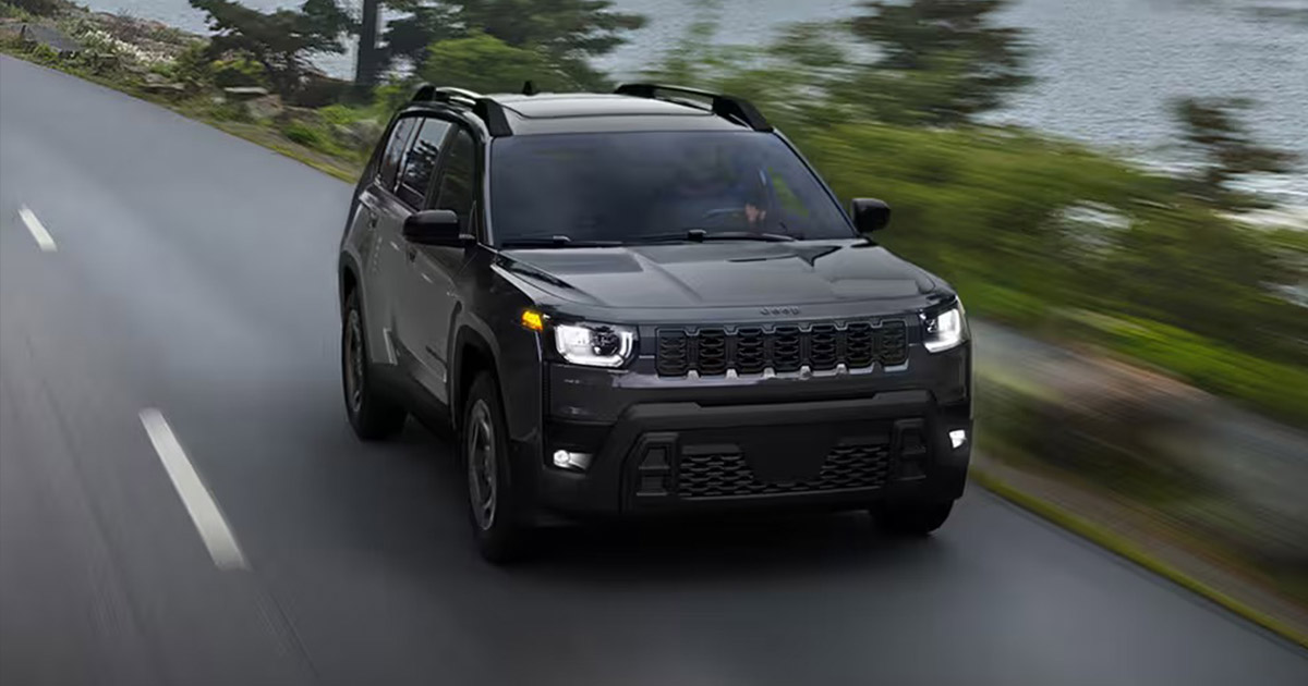 A black Jeep Cherokee driving on a scenic, wet road lined with trees, near a lake. The sky is overcast, lending a serene and adventurous mood to the scene.