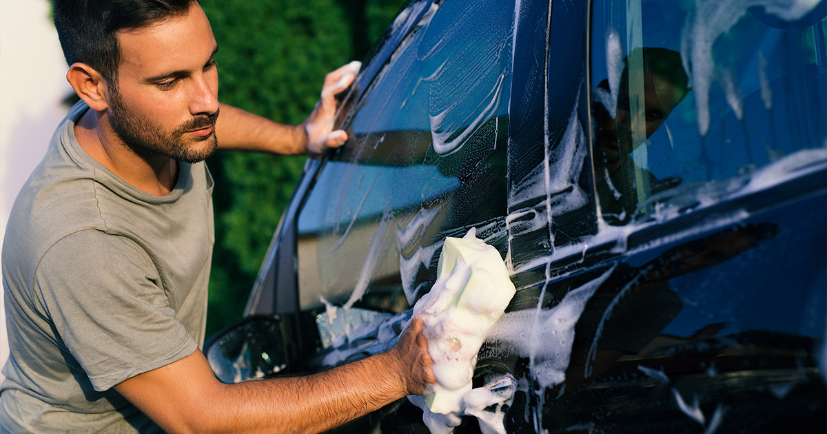 Man washes a car with a sponge, covered in soap suds, under sunlight. His expression is focused. The scene conveys cleanliness and care.