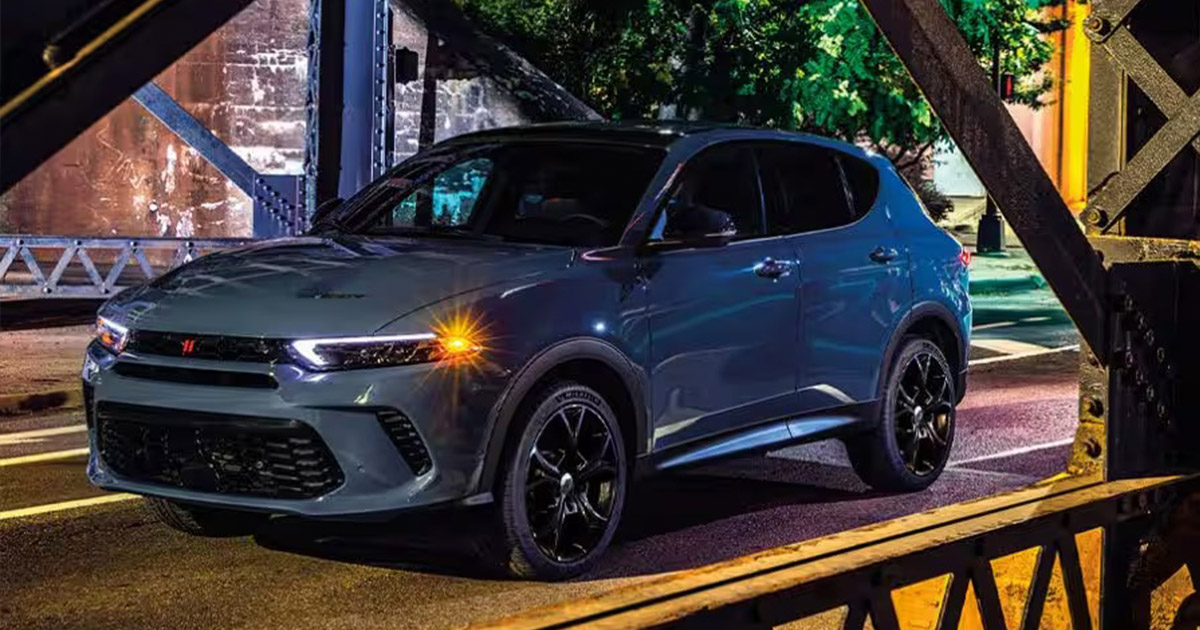 A sleek gray Dodge Hornet SUV with glowing headlights is parked under a metal bridge at night. The setting is urban, with a mix of industrial and natural elements.