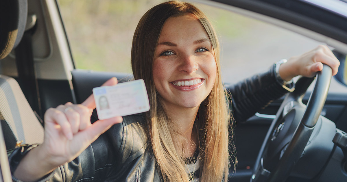 Smiling woman in a car holds up a driver's license, showcasing it proudly. She wears a leather jacket and the image conveys a sense of achievement.