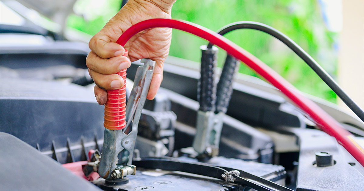 A hand attaches a red jumper cable to a car battery's terminal, set against an open hood. The scene conveys focus and urgency in car maintenance.