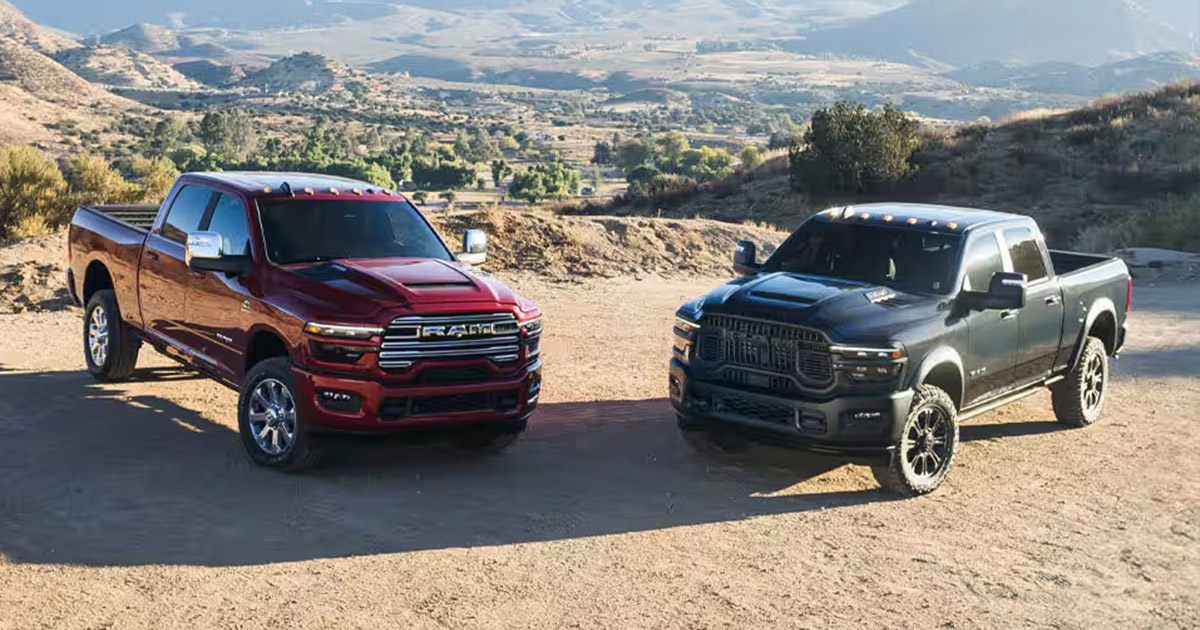 Two large RAM pickup trucks, one red and one black, are parked on a dirt path with a mountainous landscape in the background, under a clear blue sky.