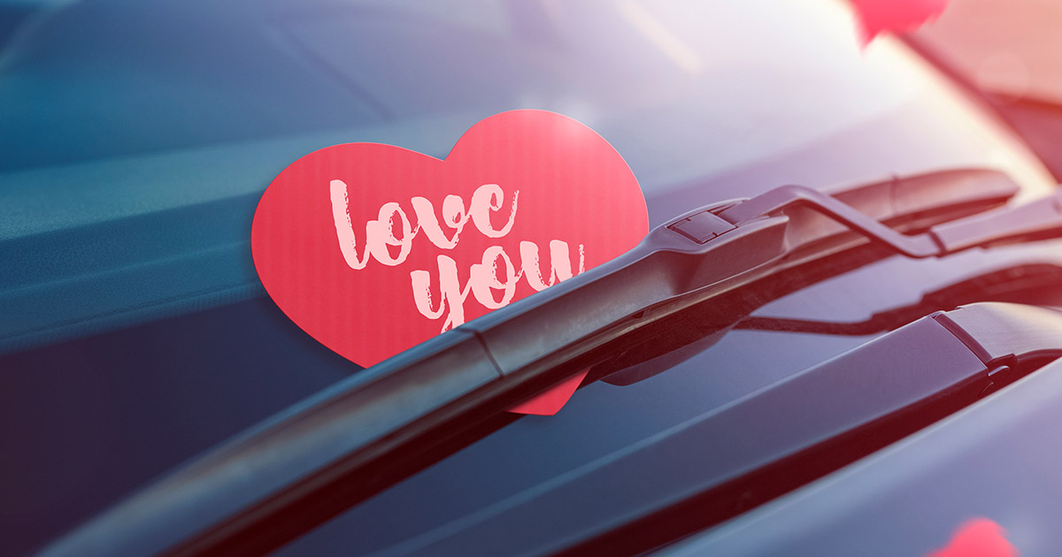 A red heart-shaped note with "love you" in white cursive is tucked under a windshield wiper on a car, conveying a warm, romantic message.